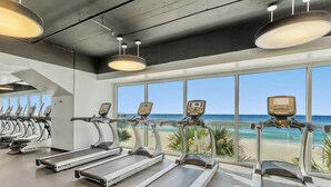 Ein Fitnessstudio mit Laufbändern und Trainingsgeräten, große Fenster mit Blick auf den Strand.