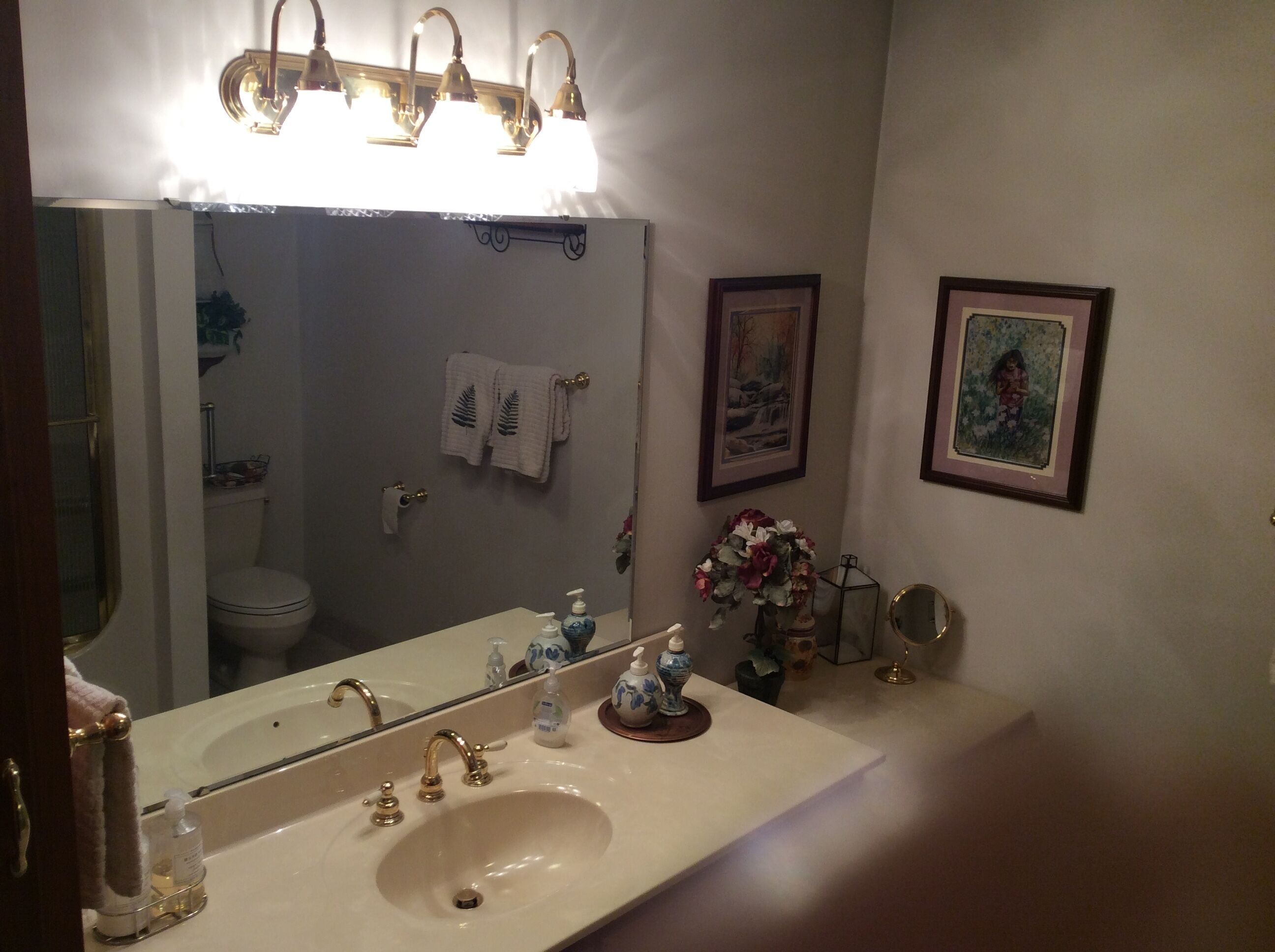A bathroom with a sink, mirror, and toilet. There are towels hanging on the wall, a framed picture, and a vase with flowers.
