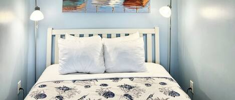 A bedroom with a bed, a ceiling fan, two bedside lamps, and a painting of boats on the wall.