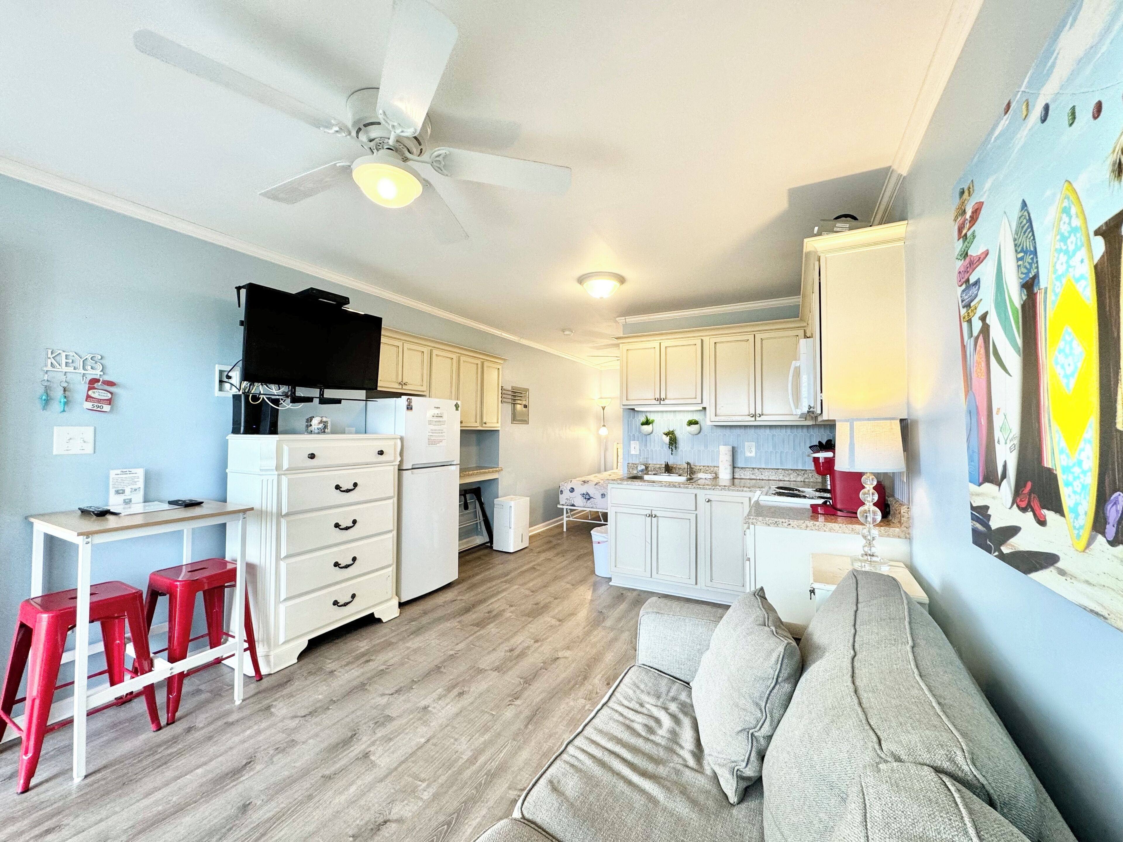 A compact living space with a kitchenette, a dining area with red stools, a sofa, and a ceiling fan.