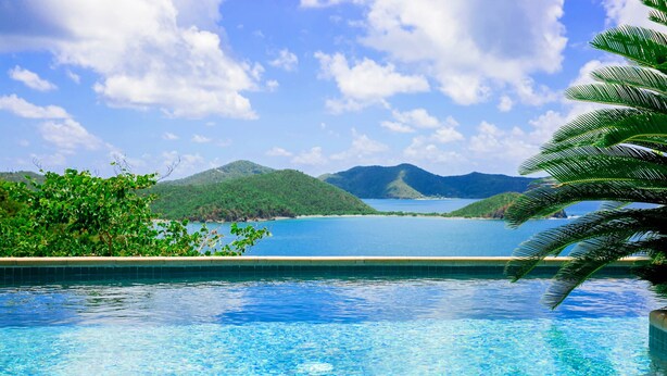 Une piscine à l’eau d’un bleu cristallin, entourée d’une végétation luxuriante et offrant une vue sur une mer calme et des montagnes lointaines.