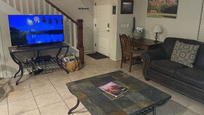 Ein Wohnzimmer mit Flachbildfernseher, Couchtisch mit Zeitschrift, Sofa, Esstisch und Treppe mit Gemälde.