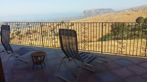 Ein Balkon mit zwei gestreiften Liegestühlen und einem kleinen Korbstuhl, mit Blick auf eine hügelige Landschaft und das Meer.