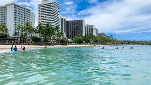 Une plage avec une eau turquoise et limpide, des palmiers et, en arrière-plan, des immeubles de grande hauteur.