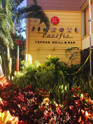 A building with a sign reading "Jinkoku Pacific Teppan Grill & Bar" surrounded by lush tropical plants.