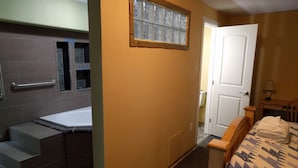 Un bagno con vasca, doccia e servizi igienici. Nell'stanza adiacente, un letto di legno con copripiumino decorato e una porta bianca.