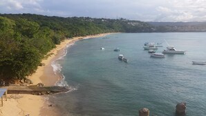 Uma praia com barcos ancorados na água, uma faixa de areia e uma encosta arborizada ao fundo.