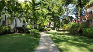 Uma trilha serpenteia por um jardim exuberante, com palmeiras e arbustos variados, em meio a edifícios.