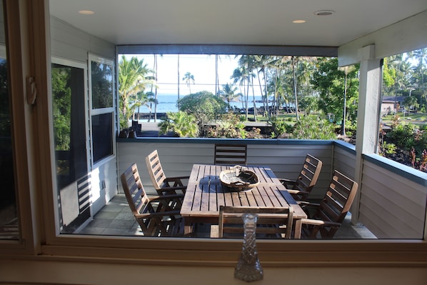 Un patio techado con una mesa y sillas de madera, con vistas a un paisaje tropical con palmeras y el océano.