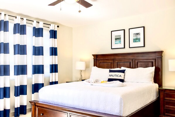 A neatly made bed with a white comforter and a pillow labeled "NAUTICA," a wooden headboard, a ceiling fan, and two framed pictures on the wall. - Montego Bay Luxury 3BR Villa Ocean Views & Pool (Montego bay)