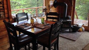 Una cabaña de madera con una mesa de comedor preparada para cuatro personas, una chimenea y un ventanal con vistas a un bosque.