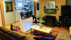 A living room with a sofa, a coffee table, and a fireplace. There is a television and a kitchen visible in the background. - Great house for making memories! Two living areas, bar, and pool table (Alpine)