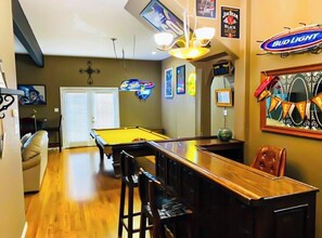 A pool table in a room with a bar, a ceiling fan, and various wall decorations including a neon sign and a Bud Light logo.