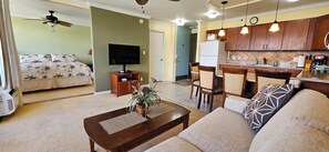 A living room with a TV, sofa, coffee table, and a dining area with chairs and a kitchen in the background.