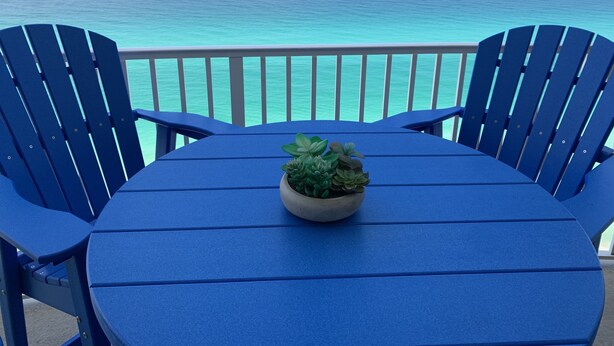 A blue table with a potted plant, surrounded by blue chairs, overlooking a turquoise sea with a distant island.