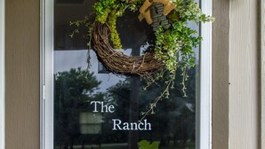 Une porte surmontée d’une guirlande, un pot de fleurs placé à côté de la porte, et l’inscription « The Ranch » sur la porte en verre.