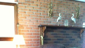 Une pièce avec une cheminée en briques, un fauteuil à capitons, une petite table avec une lampe et un chariot en métal.