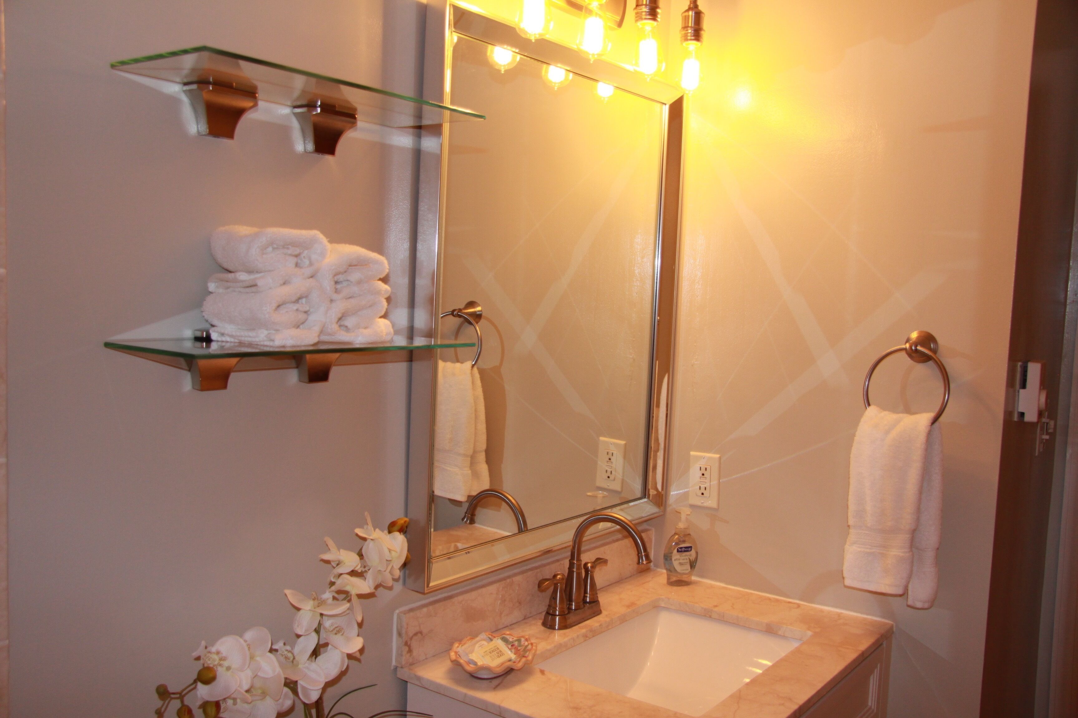 A bathroom with a glass shelf holding folded towels, a sink with a mirror above it, and a towel hanging on a ring.