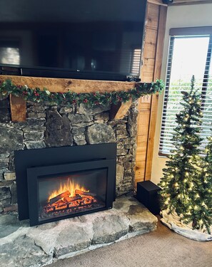 A stone fireplace with a fire, a wooden mantel, and a Christmas tree with lights.