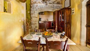 Un coin repas agrémenté d’une arche en pierre menant à la cuisine, de chaises en bois, d’une table sur laquelle se trouve un panier de fruits et une bouteille de vin.