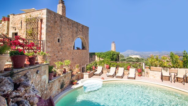 Un espace piscine avec des chaises longues et des plantes en pot, un bâtiment en pierre doté d’une arche, et une vue sur la mer.