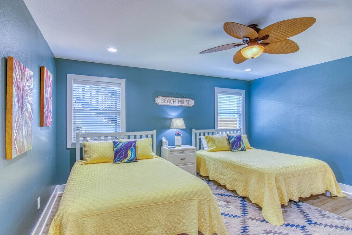 A bedroom with two beds, a ceiling fan, and a sign that reads "BEACH HOUSE".