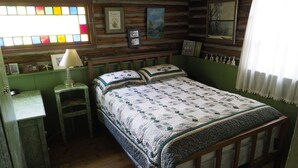 A rustic wooden cabin bedroom with a bed, a nightstand, a lamp, and a window with stained glass. - Cozy Log Cabin ON THE WATER with the Best Sand Beach in Door County! (Sturgeon Bay)