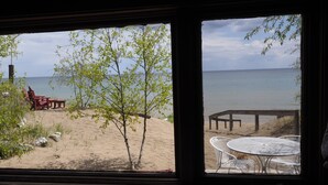 A view from a window overlooking a beach with a wooden deck and a table with chairs. - Cozy Log Cabin ON THE WATER with the Best Sand Beach in Door County! (Sturgeon Bay)