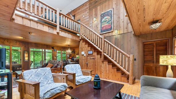 Ein geräumiges Blockhaus-Interieur mit einer Holztreppe, einer gemütlichen Sitzecke mit gemusterten Kissen und einem großen Fenster mit Blick auf den umliegenden Wald.