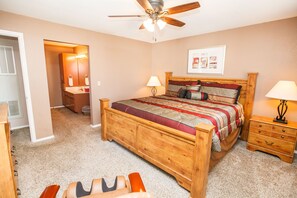 Une chambre à coucher avec un lit en bois, une table de chevet, une commode, un ventilateur de plafond et une porte donnant sur une autre pièce.