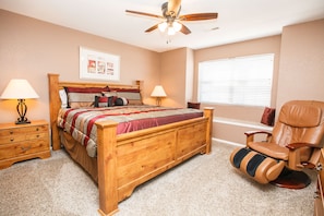 Une chambre à coucher avec un lit en bois, deux tables de chevet avec des lampes, un ventilateur de plafond, une fenêtre avec des stores et un fauteuil inclinable.