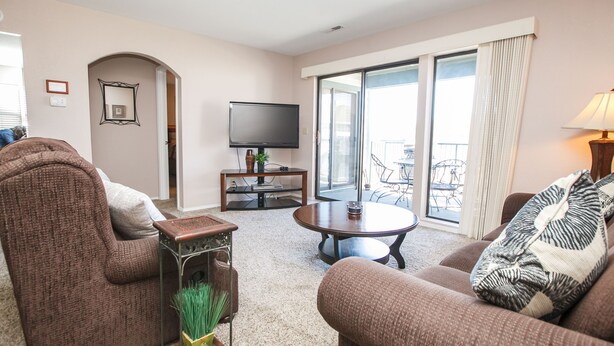 A living room with two brown sofas, a coffee table, a flat-screen TV, and a balcony with a view.