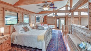 A log cabin bedroom with a large bed, a ceiling fan, and a view of the outdoors. - Mata'Zamo cliffside cabin, 1000 acres hiking trails, farm animals, family fun! (Glen Rose)