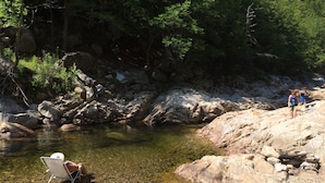 Orilla rocosa de un río con agua cristalina, una silla y dos personas caminando.
