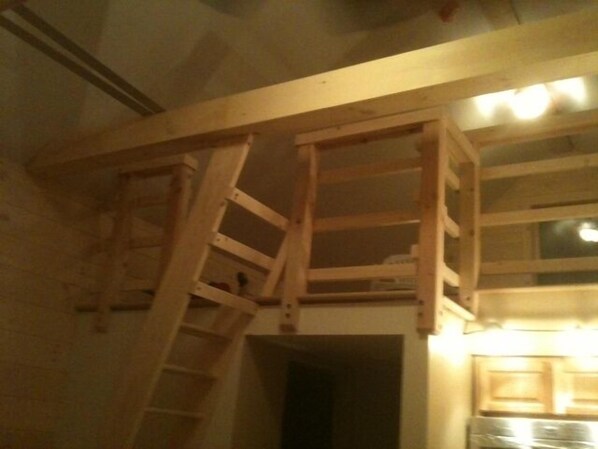 A room with a wooden loft bed structure, a ladder, and a kitchen area with cabinets.