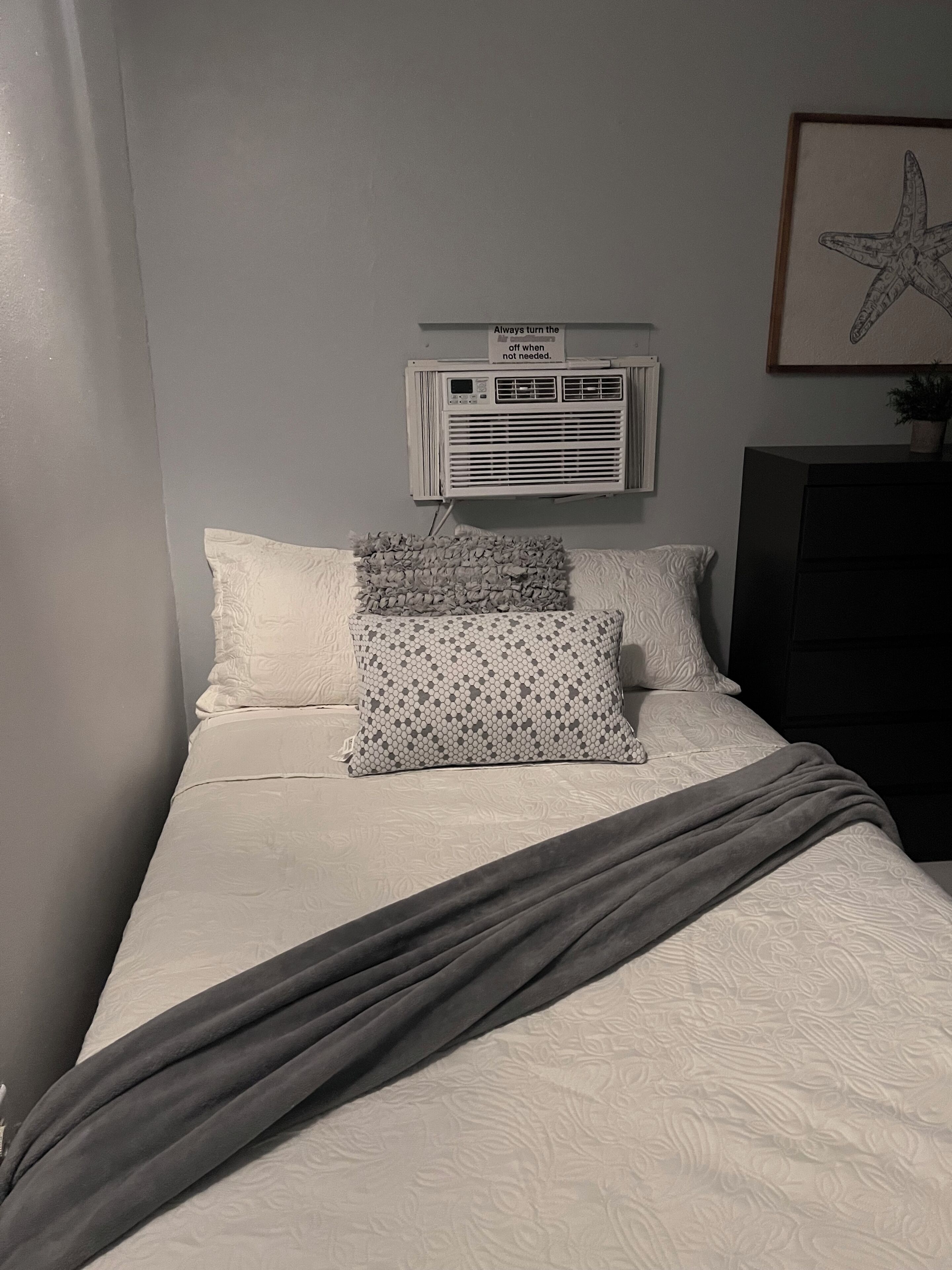 Una camera da letto con un letto, cuscini, una coperta, un condizionatore d'aria e un quadro appeso al muro.