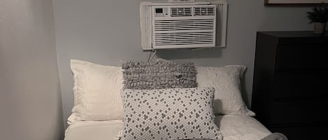 Una camera da letto con un letto, cuscini, una coperta, un condizionatore d'aria e un quadro appeso al muro.