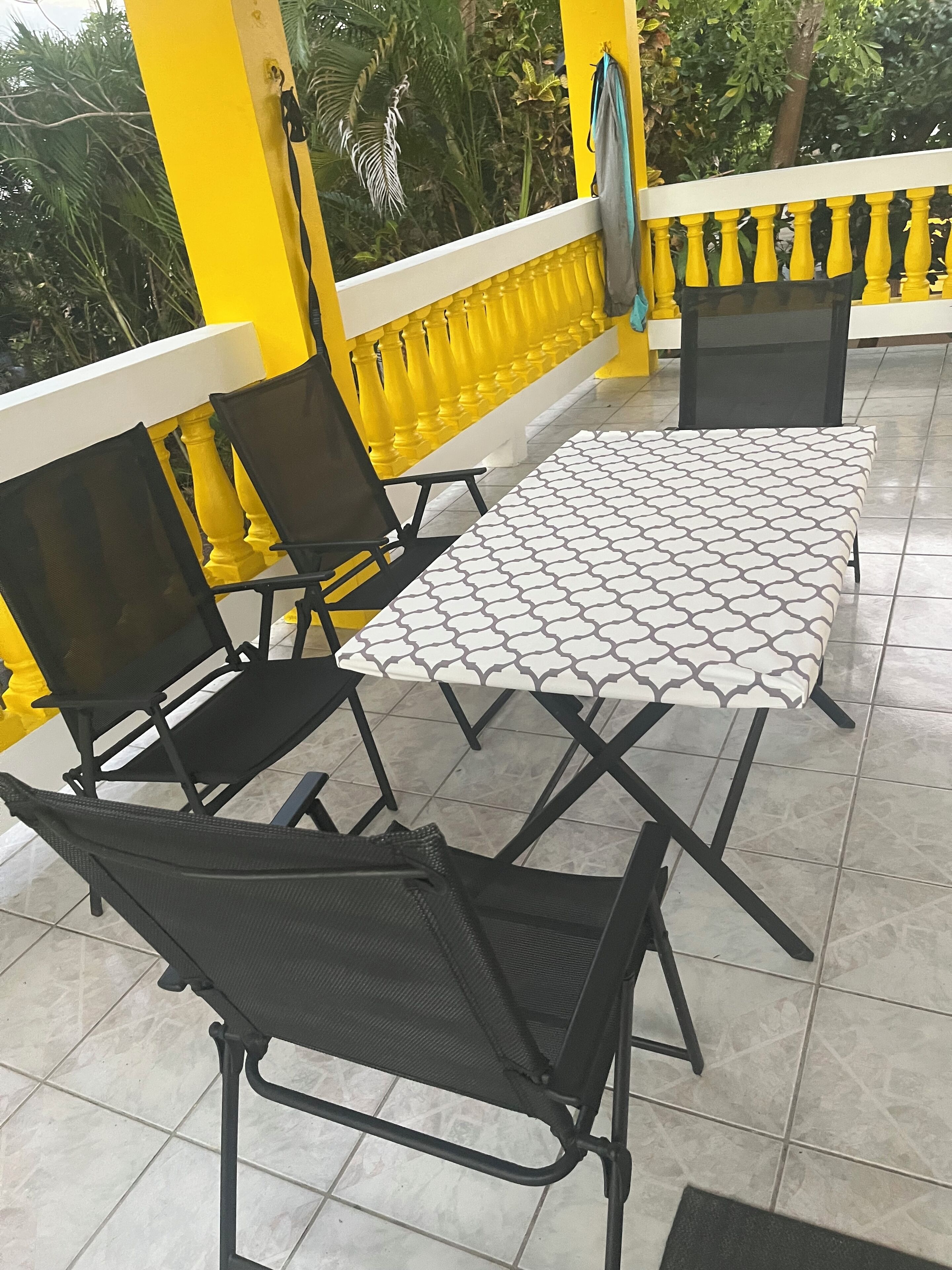 A balcony with yellow railings, a table with a patterned tablecloth, and two black chairs.