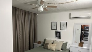 A bedroom with a bed, a ceiling fan, an air conditioning unit, and a small side table.