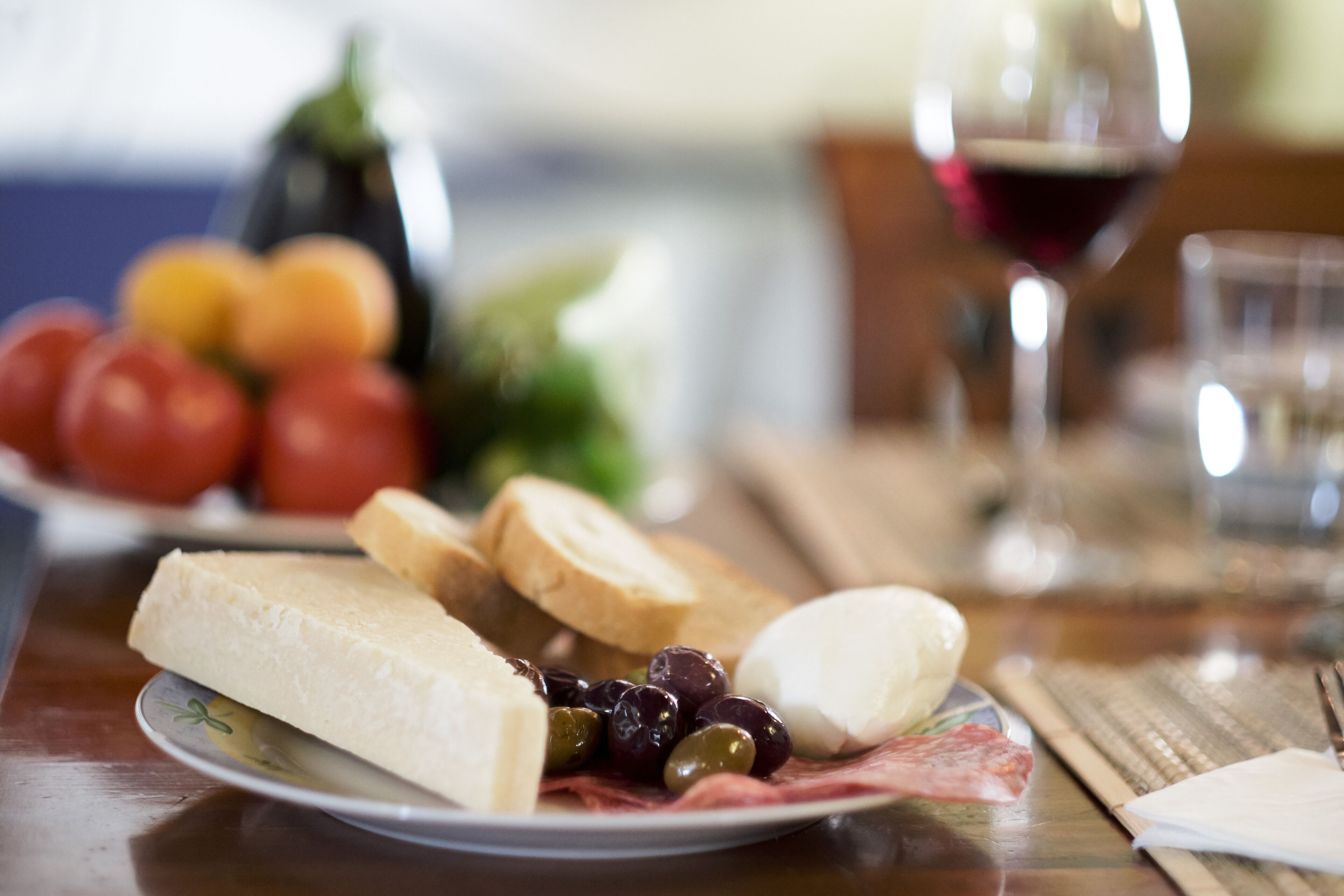 Ein Teller mit Käse, Oliven und Brot auf einem Tisch, im Hintergrund ein Glas Rotwein und eine Schale mit Obst.