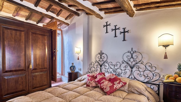 Un dormitorio con un amplio armario de madera, una cama con ropa de cama estampada, una pared decorativa con una cruz y una mesita de noche con fruta.