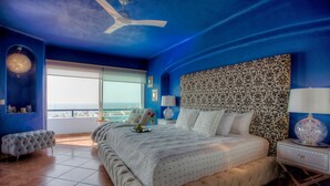 A bedroom with a large bed, a ceiling fan, a view of the ocean, and a patterned headboard.