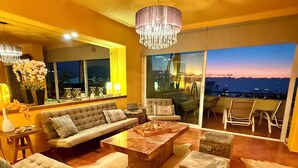 A modern living room with a sofa, coffee table, ottoman, and chairs. There is a chandelier, a window with a city view, and a balcony with chairs.