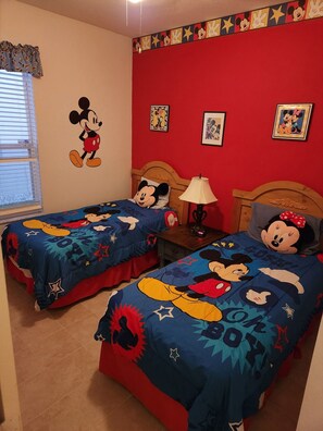 Habitación con dos camas, cada una con ropa de cama temática de Mickey Mouse. Decoración de pared que incluye a Mickey Mouse y Minnie Mouse, y una ventana con persianas.