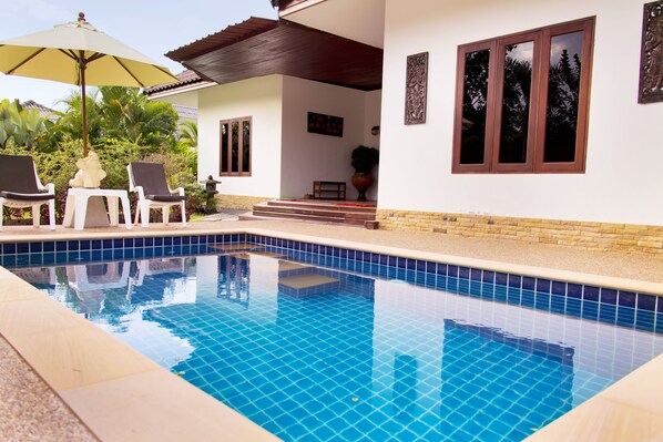A swimming pool with tiled edges, two lounge chairs, a table, and an umbrella.