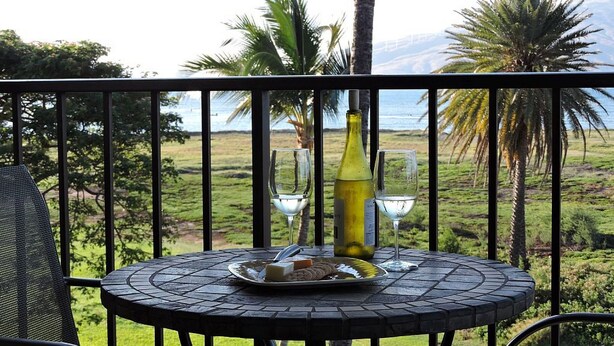 Een ronde tafel met een fles en twee glazen op een balkon met uitzicht op een grasachtig landschap met palmbomen en de zee.