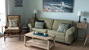 A living room with a sofa, armchair, coffee table, and a painting of the ocean. - 19th floor with  beach view free beach service and 10% weekly discount (Panama City Beach)