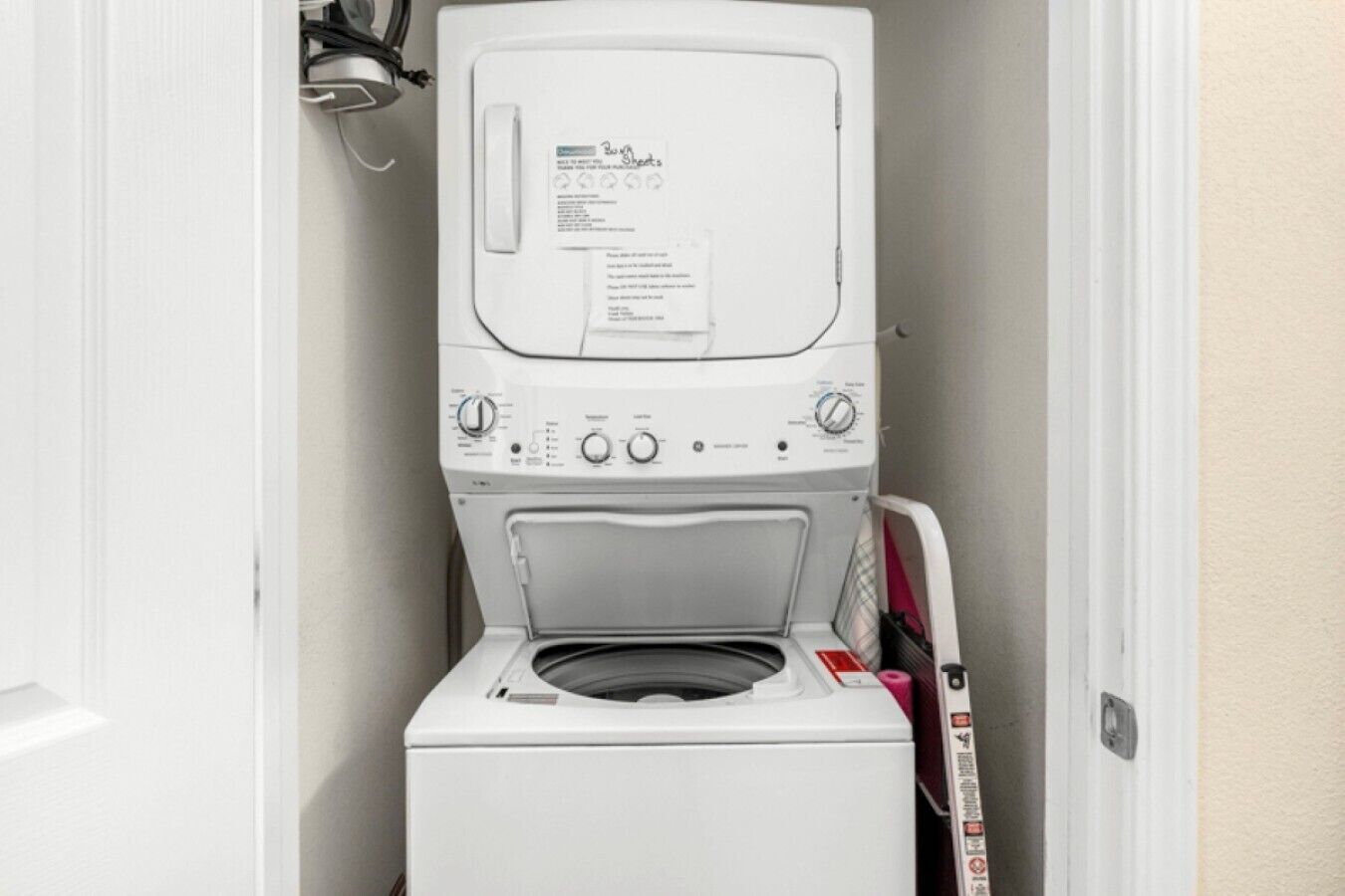 Une petite machine à laver blanche à chargement frontal, installée dans un petit espace buanderie, avec une porte d’un côté et un sèche-linge fixé au mur de l’autre.