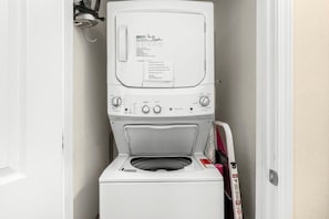 A compact, white front-loading washing machine in a small laundry room with a door on one side and a wall-mounted dryer on the other.