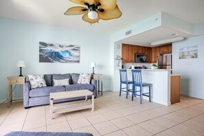 Ein Wohnzimmer mit Sofa, Couchtisch und Deckenventilator. Es gibt eine Küchenzeile mit Mikrowelle, Kühlschrank und einer Theke mit zwei Stühlen.